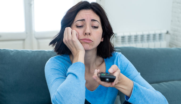 Young upset woman on sofa using control remote zapping bored of bad TV shows and programing . Looking disinterested, aloof and sleepless. People, too much bad television and Sedentary lifestyle.